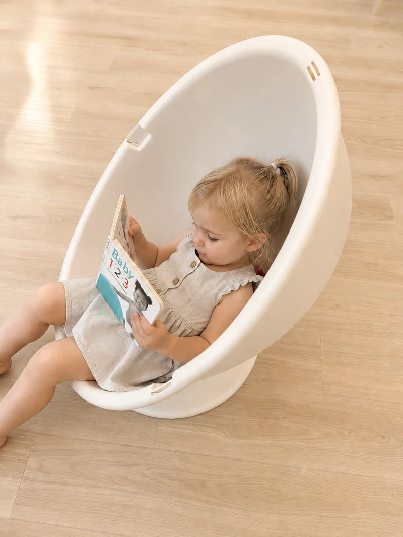 A toddler curled up in the white egg chair with a board book