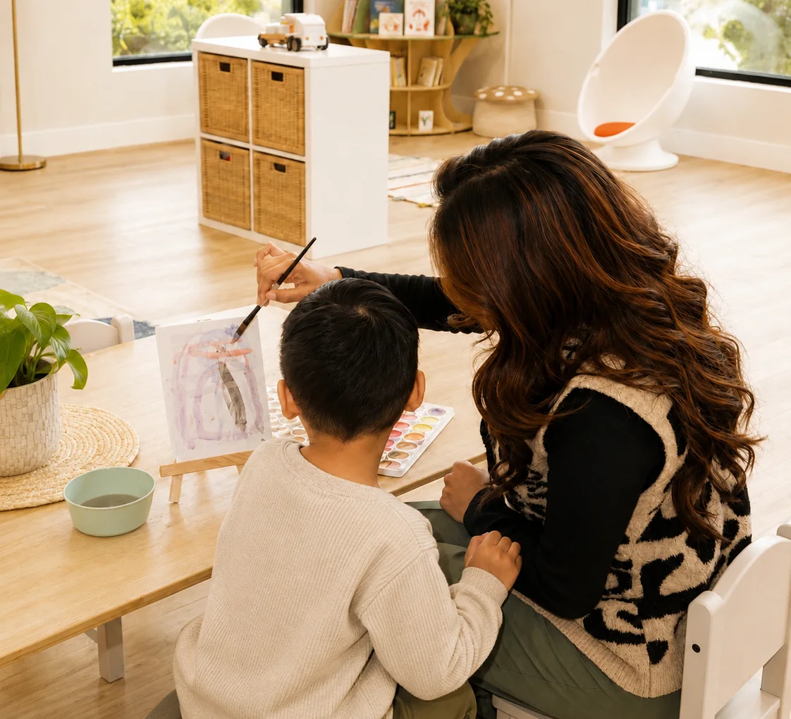 An educator guiding a child&rsquo;s watercolour painting at a low table, the reading corner and egg chair behind them
